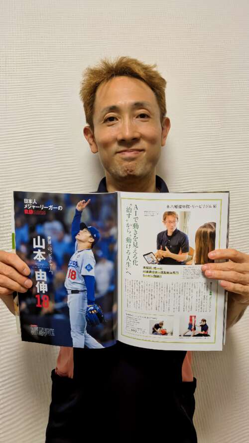 男性が雑誌を手に持ち、笑顔でカメラに向かって立っている。雑誌にはメジャーリーガーの写真と、AI姿勢分析を使ったリハビリ指導の紹介記事が掲載されている。