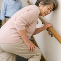 手すりを握り、膝を押さえながら階段を上がる高齢女性。後方で見守る男性の姿もある。