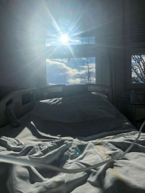 窓際のベッドに一人で座り、外を静かに見つめる高齢者の後ろ姿。光が差し込む室内は静かで、孤独と時間の長さを感じさせる。